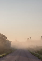 Grey sky fades into peachy mist that covers an old country road receding over a small rise in the distance.